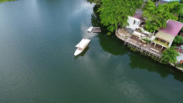This drone footage shows a white boat on the calm waters of Lake Tondano, Minahasa, North Sulawesi, with a lakeside building surrounded by lush greenery.