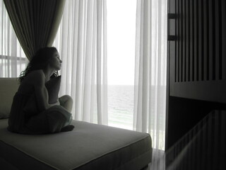 A girl with dark curly hair is sitting on the couch and looking out the window at the ocean. the...