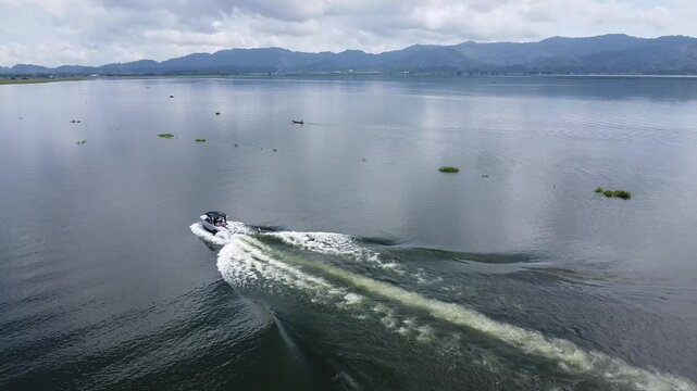 A speedboat glides across Lake Tondano, Minahasa, North Sulawesi, leaving waves on the calm water. In the distance, a small boat floats, with green mountains and a cloudy sky in the background.