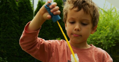Little cute boy in brightly coloured T-shirt uses long wand for blowing huge bubbles. Child enjoys...