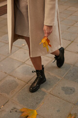 Close up of woman's legs in beige coat and black boots with yellow maple leaf. Photo fashion walks