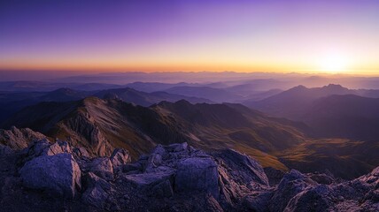 Naklejka premium A panoramic view from the summit of a majestic mountain at golden hour. The sky is a blend of warm golden and purple tones, and the vast expanse of valleys and distant