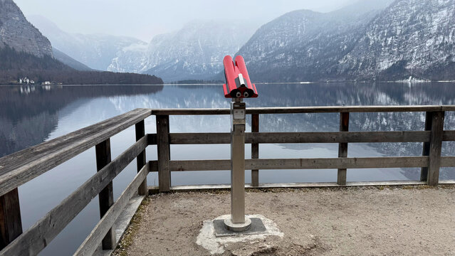 Red binoculars on viewing platform by tranquil lake Hallstatt,Austria. surrounded by snowcapped Alps highlight the serenity of nature in winter. Frosty scene exudes calmness and peaceful contemplation
