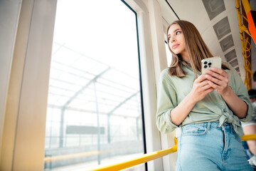 Young woman enjoying a sunny day on urban public transport holding a smartphone while gazing out...