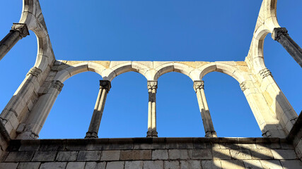 Arched ruins of an ancient structure against a clear blue sky. The stone columns with intricately carved capitals evoke a sense of antiquity and history