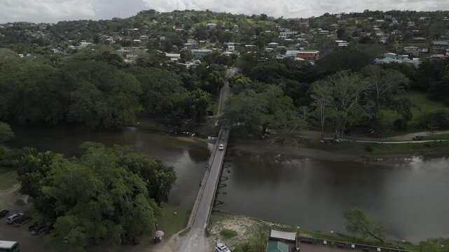 Drone facing east flies to the right along the Macal River lined with trees leading up to metal suspension Hawkesworth Bridge in San Ignacio, Belize on partially cloudy afternoon