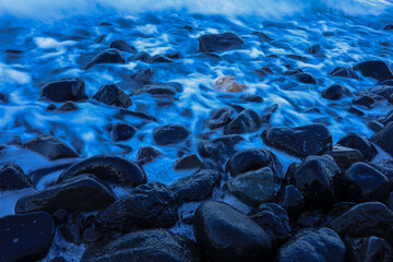 Smooth black volcanic rocks, with a single lighter-colored stone standing out among them, as misty ocean waves wash over. Captured with long exposure for a soft, ethereal effect.