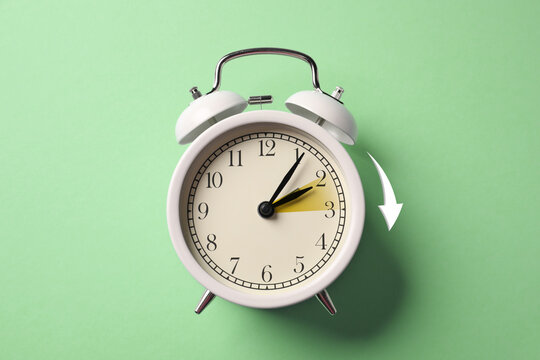 Daylight saving time. White alarm clock on light green background, top view. Highlighted area and arrow showing time change by one hour forward - Powered by Adobe