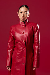 Fashionable asian brunette in a red leather coat poses confidently with a smile against a matching red background