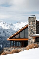This contemporary stone house features expansive windows that offer stunning views of the snowy mountains, set against a picturesque winter landscape drenched in bright daylight
