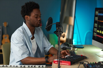 Young Black man sitting at desk playing electronic keyboard in home recording studio while adjusting sound settings using computer monitor