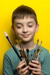 Smiling child in teal shirt holding colorful paintbrushes, yellow background. Perfect for themes of creativity, childhood art, playfulness, and joyful expression.