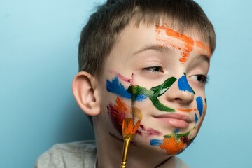 Child with colorful face paint holding a paintbrush, blue background. Perfect for themes of creativity, childhood art, playfulness, and artistic exploration