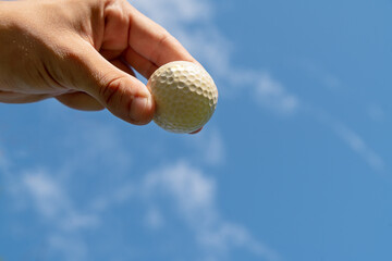 Hand holding golf ball against beautiful blue sky with white clouds.