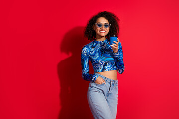 Trendy young woman in vibrant outfit holding a cup against a bold red background, embodying style,...