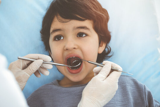 Cute arab boy sitting at dental chair with open mouth during oral checking up with doctor. Visiting sunny dentist office