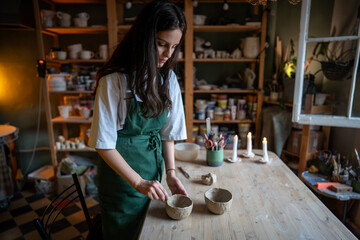 Creating handicraft pottery. Working table in ceramics studio with woman master shaping wet clay cup. Female potter sculpting decorating handmade bowl in workshop. Art, small business concept