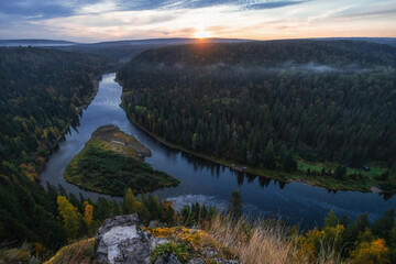 Fototapeta premium A stunning sunset casts golden hues over a winding river nestled between dense forests and hills, showcasing the beauty of untouched nature during twilight hours