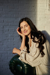 young woman with long dark hair rests her chin on her hand while seated in a modern, bright room. Soft natural light illuminates her friendly expression, creating a warm atmosphere