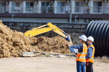 supervisor standing at a construction site, reviewing blueprints. A large pile of dirt and an...