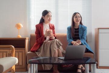 Two asian businesswomen are sitting on a sofa, discussing during a business meeting while using a laptop placed on a table in front of them, in a modern office with a large window