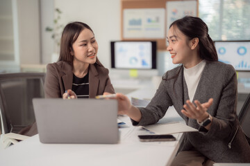 Two professional businesswomen engage in a collaborative discussion at a modern office. They use a laptop and documents to share ideas, enhancing productivity and teamwork