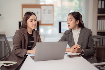 Obraz premium Two Asian businesswomen sitting at a modern office table, using a laptop while engaging in a focused discussion about work during a productive meeting