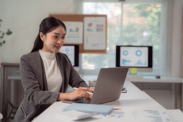 Young businesswoman in a modern office setting, focused on her laptop. Charts and graphs are visible on screens and a bulletin board, indicating a productive work environment