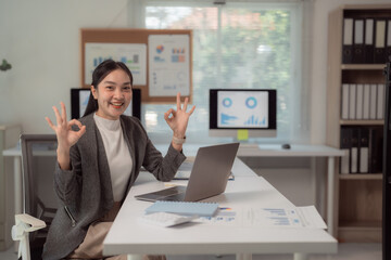 Confident businesswoman showing success gesture while sitting at desk in a modern office. Multiple charts and graphs displayed on screens, emphasizing data-driven decision making