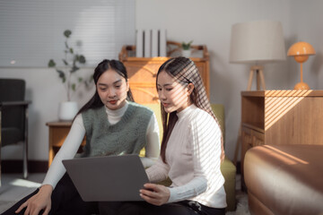 Two asian businesswomen are working from home using laptop sitting on floor in living room, blinds casting shadows on them and the furniture