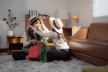 Two happy female friends are sitting on a carpet in the living room, trying on hats after a shopping spree, surrounded by colorful shopping bags, enjoying their time together