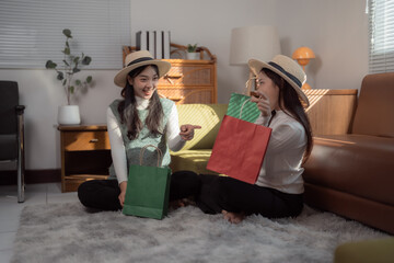Two happy female friends wearing summer hats are sitting on the floor at home, showing shopping...