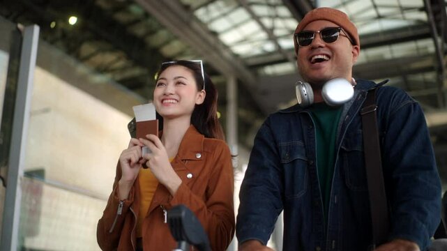 Happy asian couple walking through the airport terminal with luggage, using a smartphone at the boarding gate to checking time flight and ready for their vacation trip.
