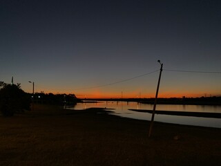 A calm river at sunset, reflecting the orange sky, with a leaning pole and bridge in the distance