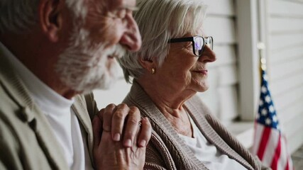 Senior couple standing closely together, holding hands lovingly while gazing proudly at waving american flag, sharing tender moment of patriotic connection on home porch during peaceful daylight - Powered by Adobe