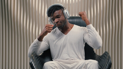 Futurism African American man male guy in white futuristic room indoors cyberspace modern technology in wireless headphones listening music listen song audio relaxing in chair dancing enjoy sound © Yuliia