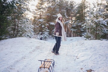 Happy young woman pulling the sled uphill in forest during walk in winter day.