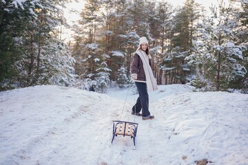 Happy young woman pulling the sled uphill in forest during walk in winter day.
