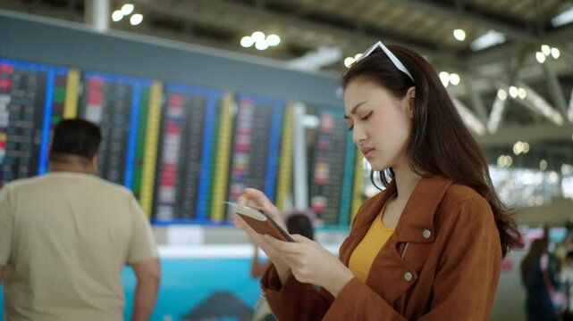 Unhappy young asian woman traveler checking at flight information schedule board while holding boarding pass at the international airport terminal, female tourist worried about missing her flight.
