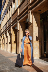 Stylish Woman Standing City Street In Fashionable outfit, Wearing Long Beige Silk Dress, Blue Cape Coat, Carrying Black tote bag, Sunglasses And Trendy ballet shoes