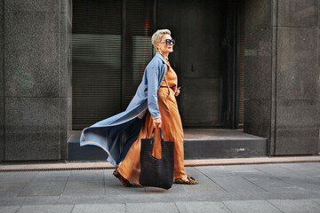Stylish Mature Woman Walking City Street In Fashionable outfit, Wearing Long Beige Silk Dress, Blue Cape Coat, Carrying Large Black Handbag, Sunglasses And Trendy ballet flats shoes