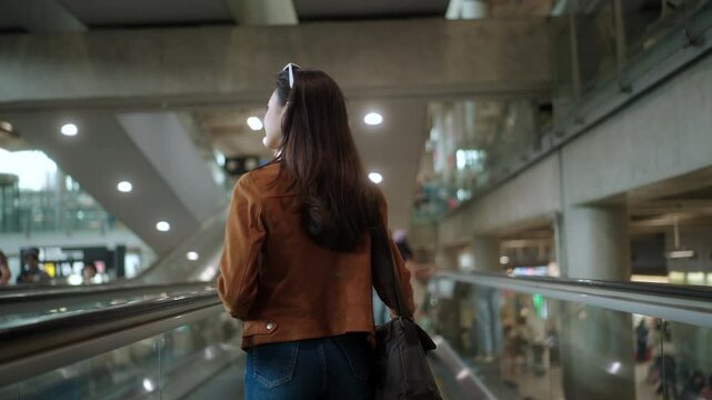 Back view of young asian woman traveler walking on escalator with holding smartphone, passport and boarding pass in the international airport terminal getting ready for vacation trip.
