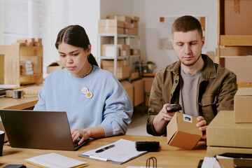 Colleagues working together in an office, with one using laptop while the other scans box, workspace features modern furniture and organized shelves in background