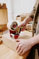 Persons hands using handheld dispenser for cutting tape on cardboard box in office setting, boxes...
