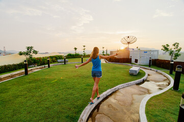 A woman in casual attire walks along a curved path on a grassy rooftop garden at sunset.