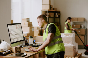 Man in high-visibility vest working on computer in warehouse surrounded by boxes and equipment,...