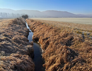 A narrow stream flows through a dry, grassy landscape towards distant mountains under a clear sky. The brown, withered grass dominates the foreground, while a lighter, barren field stretches to the ri