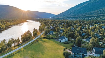Naklejka premium Aerial view of a serene river valley town at sunset, showcasing houses, green landscape, and mountains.