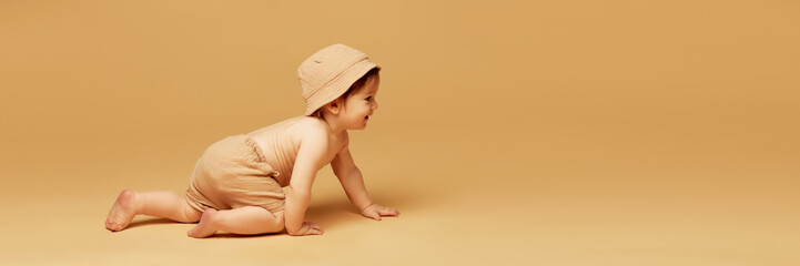 Banner. Laughing baby crawling forward wearing soft cap and shorts against warm beige background...