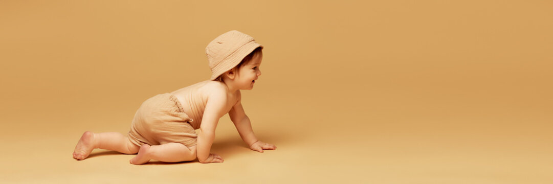 Banner. Laughing baby crawling forward wearing soft cap and shorts against warm beige background with negative space to insert text. Concept of adventure, growth, curiosity, early childhood.